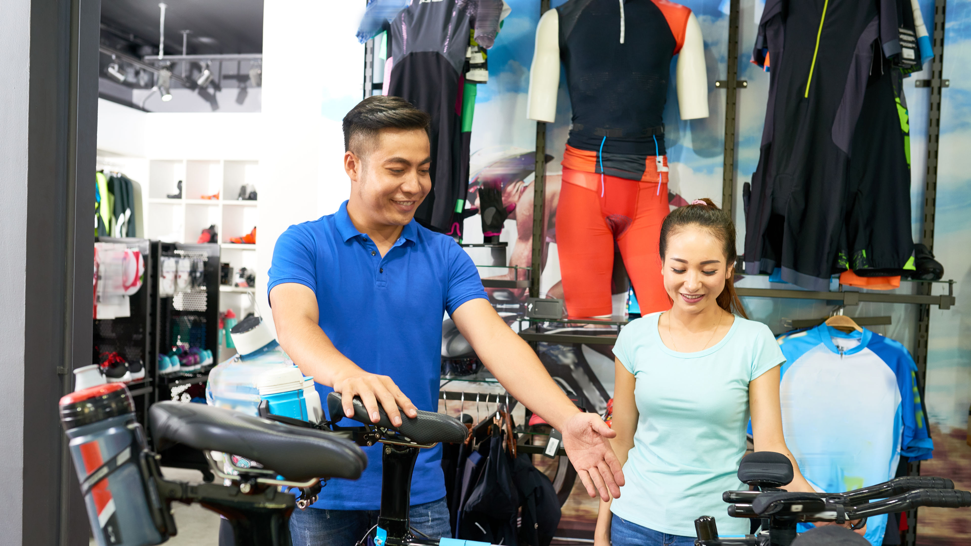 A store employee helps a smiling customer as they look at a bicycle inside a sports store.