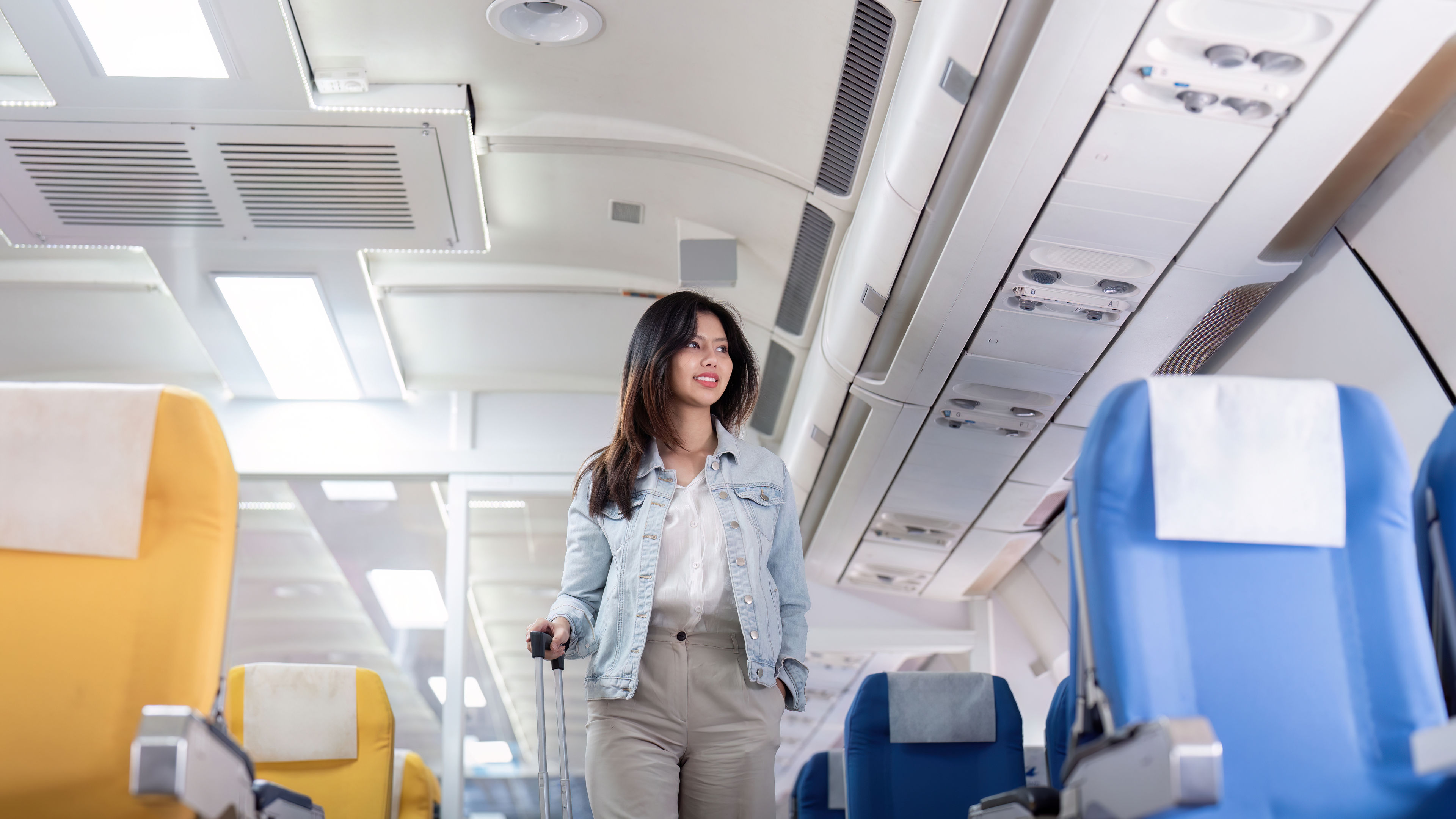 A young woman with a suitcase walks down the aisle of an airplane, excited for her trip.  