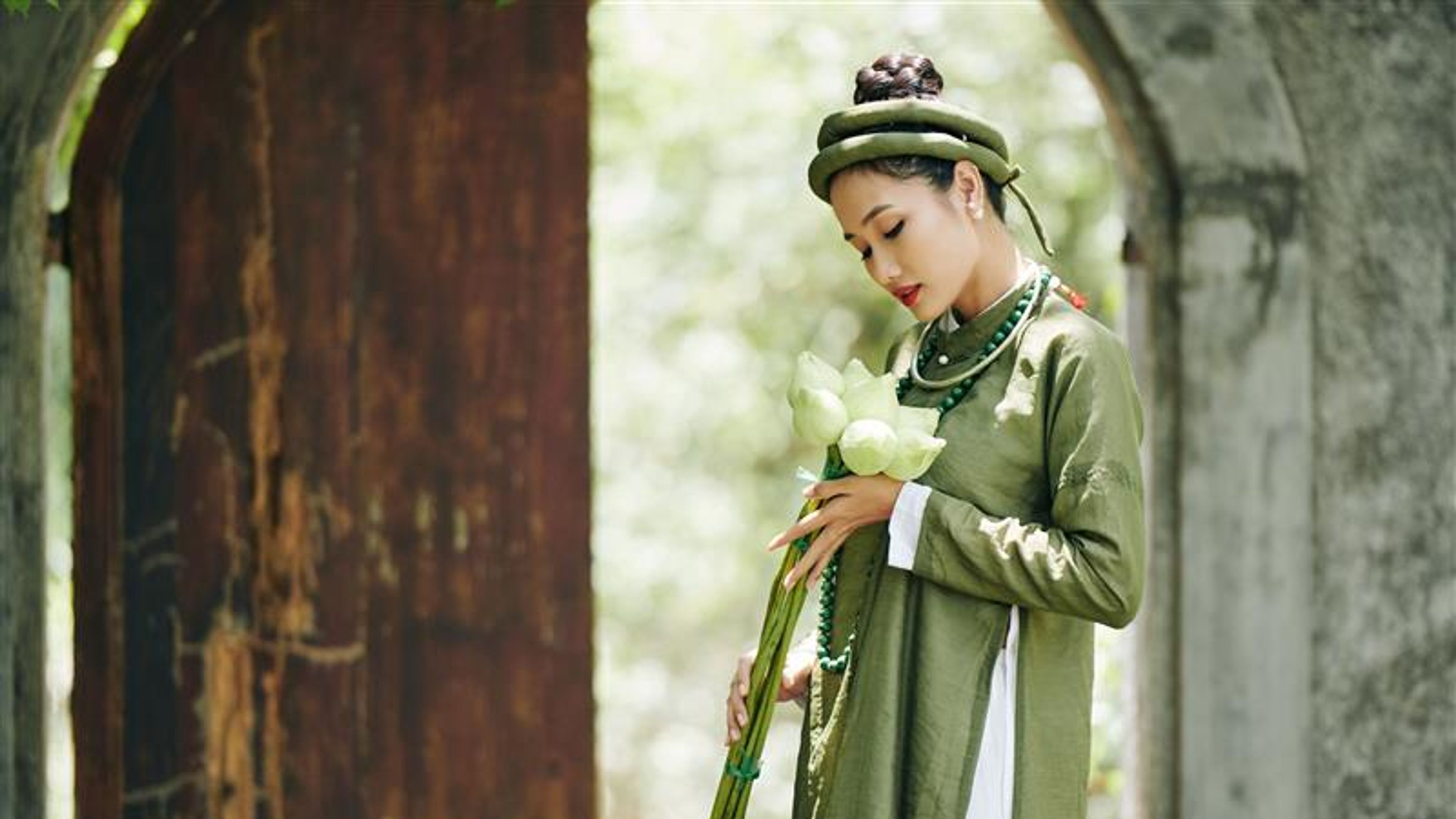 Attractive young Asian woman in traditional ao dai dress enjoying beauty of fresh lotus flowers in her hands 