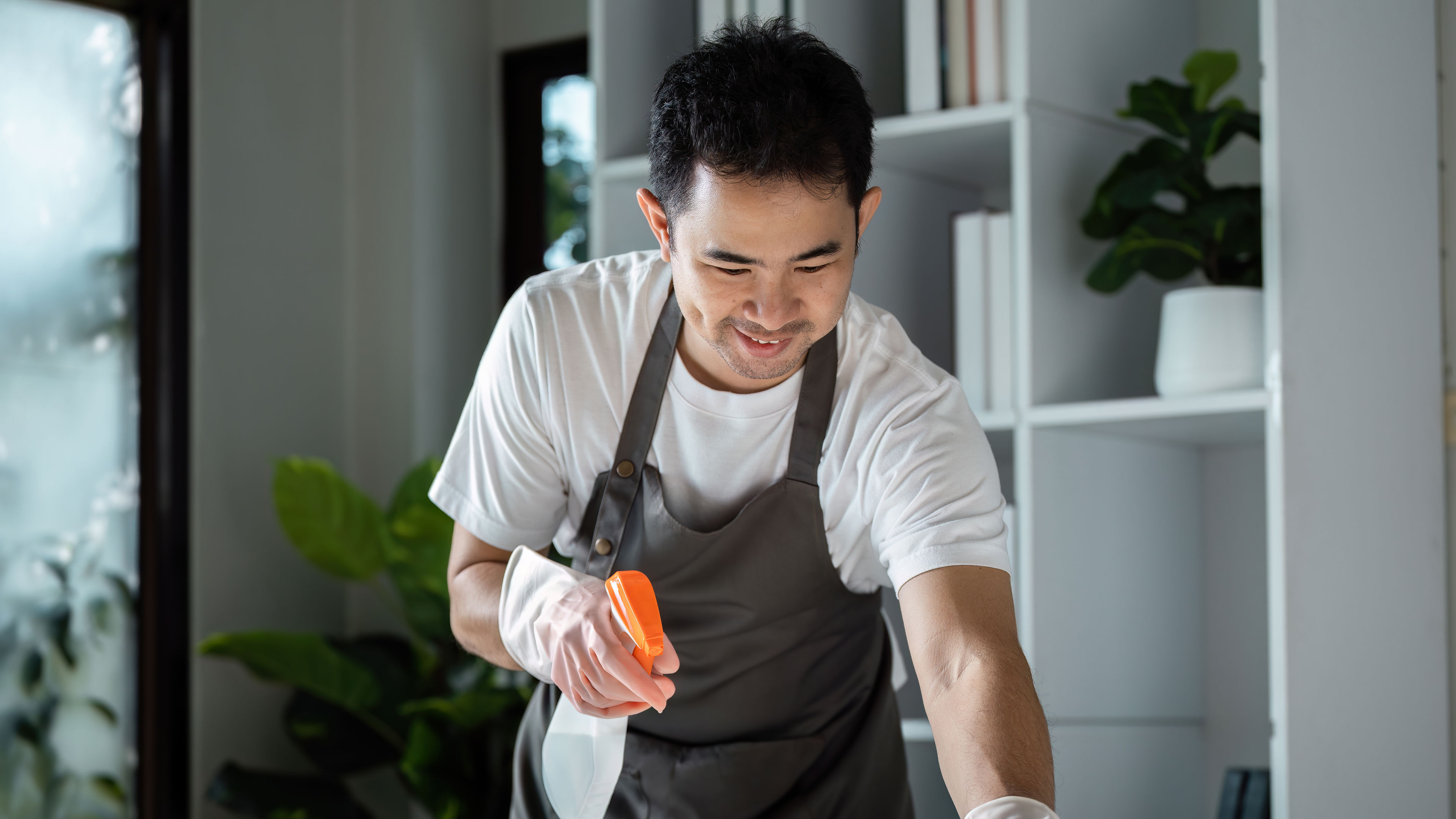 Man Cleaning Modern Home Interior with Spray Bottle and Cloth in Bright Living Room.