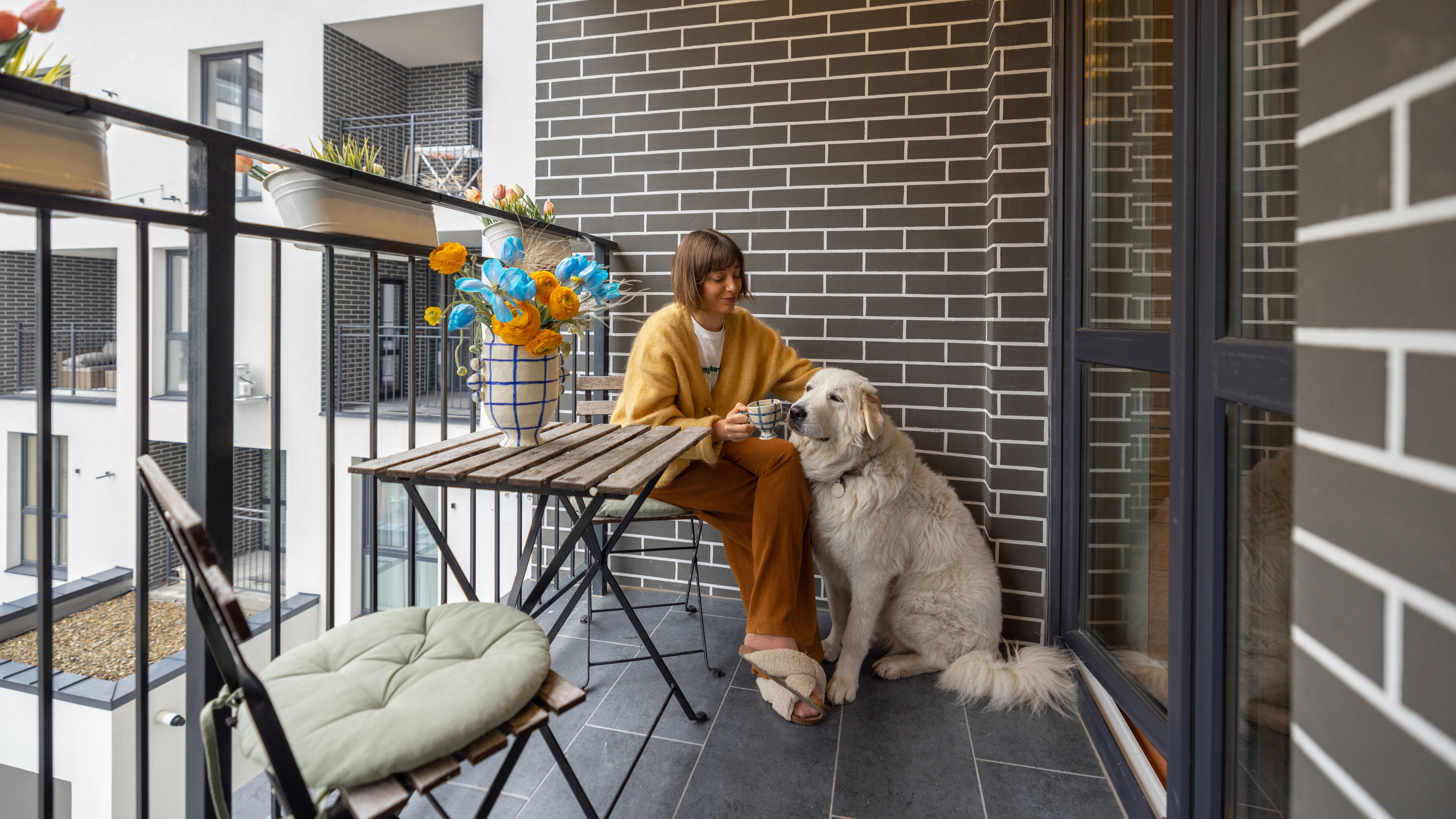 Young woman sits with her dog on balcony, enjoying coffee drink at her property during morning time. 